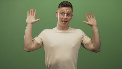 Fototapeta premium Man raising hands with palms up, smiling in beige tshirt and glasses against a green screen studio backdrop; confidence friendliness.