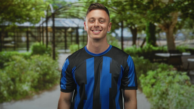Man wearing blue and black soccer jersey smiles and tilts head on a tree lined street; confidence team spirit.