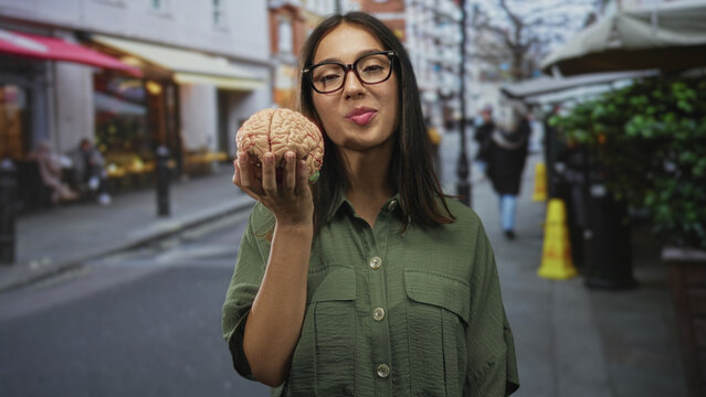 Hispanic woman in glasses holds a brain model in one hand on a busy city street, smiling and offering it toward camera; curiosity insight.