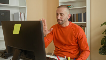 Man at computer waves hand to monitor while seated by bookshelves in building office with orange sweater visible  friendly interaction. © Krakenimages.com