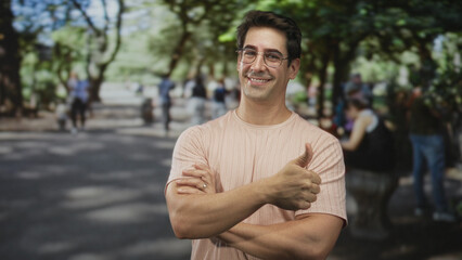 Fototapeta premium Man smiling with arms crossed and right hand giving thumbs up, wearing pink t shirt and glasses in street park with benches and trees visible; contentment.