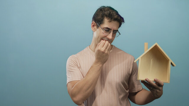 Man holding wooden house model with left hand and biting fingernails with right hand, wearing glasses and a casual shirt in studio; anxiety uncertainty.