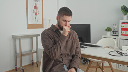 Fototapeta premium Man clutching chest seated at a desk with stethoscope in a medical building, coughing and wincing; anxiety illness.