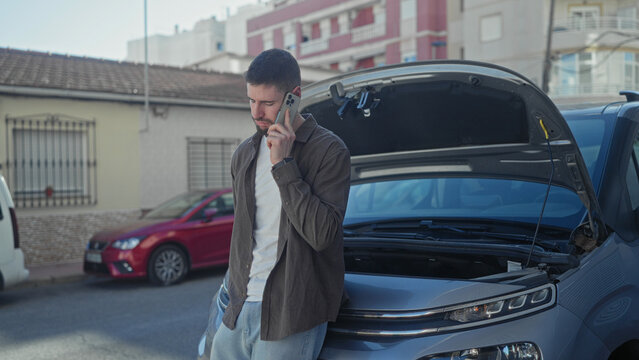 Man holding phone to ear beside car with open hood on street, leaning on bumper and looking up; roadside vehicle trouble concern.