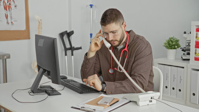 Man doctor with stethoscope holds telephone to ear and types at computer in building; focused care coordination.
