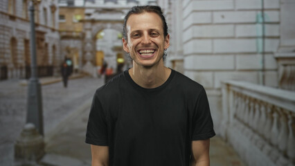 Obraz premium Man smiling with visible teeth and long hair tied back on city street beside stone balustrade and lamppost while wearing black t shirt; joy confidence.