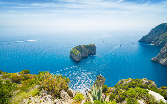 Scoglio del Monacone rock surrounded by deep blue Mediterranean water, located off southern coast of Capri, Italy, behind Faraglioni rocks. Green shrubs and agave plants grow on foreground cliffs 