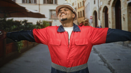 Fototapeta premium Man construction worker wearing a hard hat and red safety jacket, beard visible, arms outstretched with open palms while standing on a cobblestone street, smiling; pride duty.