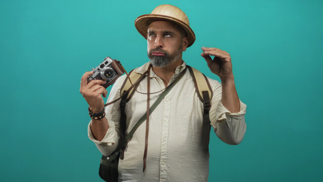 Middle aged man with grey beard holding vintage camera, wearing backpack and pith helmet in turquoise studio; playful travel adventure.