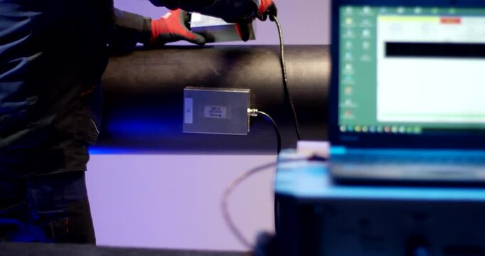 Technician in a blue uniform and gloves connects a cable to a sensor on a large black pipe, with a laptop in the foreground.