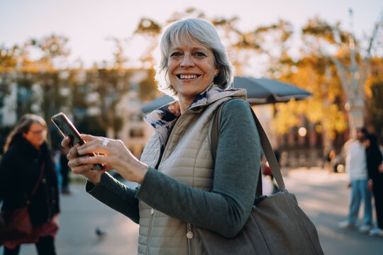Confident senior woman holding phone and smiling during street walk on sunny afternoon, reflecting empowerment and comfort through mobile technology.
