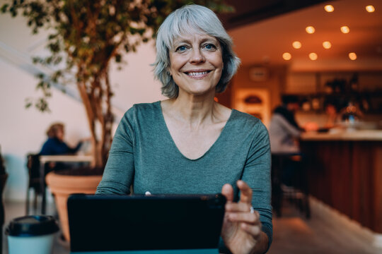 Smiling gray-haired woman using tablet at caf&eacute; table, enjoying screen interaction. Digital fluency and ease with technology define active, confident senior living.