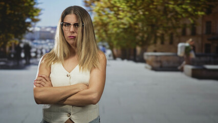 Obraz premium Woman with glasses and arms crossed, frowning on a sunlit street plaza; annoyance boundary assertiveness.