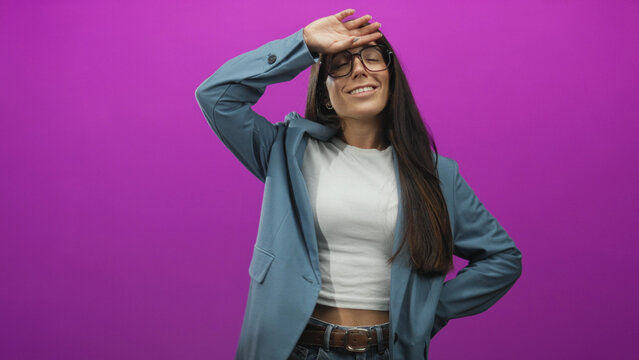 Woman with glasses shields forehead with hand, smiling and showing a casual midriff in a purple studio backdrop wearing blazer and jeans; confidence optimism.