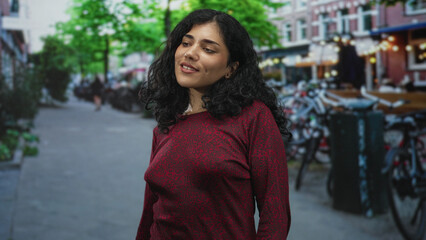 Fototapeta premium Young brunette woman smiling with face turned to side, wearing red sweater, standing near parked bicycles on a city street; serenity connection.