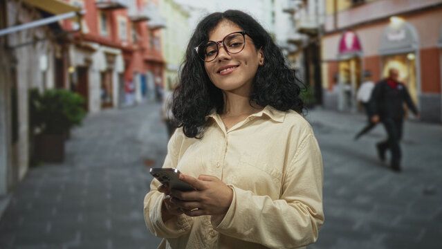 Young brunette woman texting on smartphone with both hands visible while walking on a cobbled city street lined with shops; contentment connection.