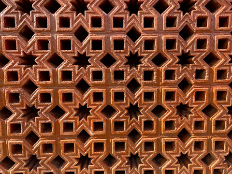 A decorative terracotta or reddish brown wall design (musharabiya or roster) featuring complex, repeating geometric patterns.