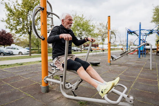Senior man with white hair and beard exercising on a rowing machine at an outdoor public gym in a park setting.