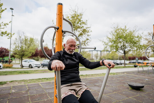 Senior man with white hair and beard exercising on a rowing machine at an outdoor public gym in a park setting.