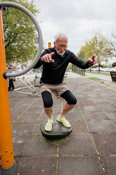 An active senior man wearing glasses and sportswear practices balance on a specialized training platform at an open-air gym facility.