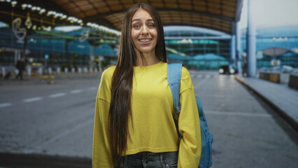 Fototapeta premium Young woman with blue backpack in yellow sweater standing at airport curb shifting from smile to pout; disappointment travel fatigue.