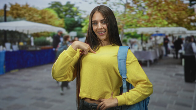 Teen girl student wearing yellow sweater and backpack points finger to chest at a bustling street market; youthful confidence.