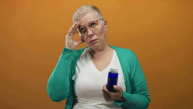 Middle aged woman holding a blue medicine bottle, looking upward with hand to temple in orange studio; confusion.