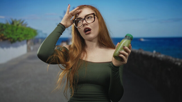 Redhead woman holding a green juice bottle with hand on forehead, shoulder exposed, making a reluctant sip gesture on a street; indifference.