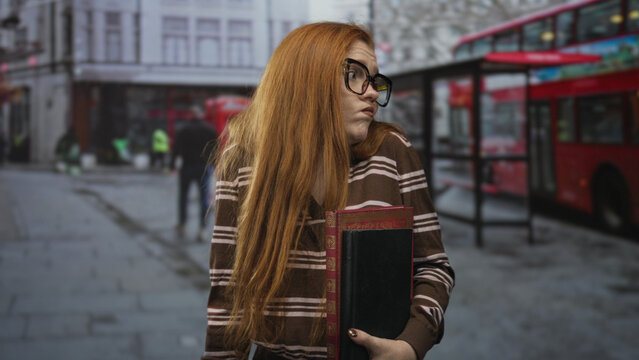 Redhead woman holding books and shrugging with an open hand on a city street beside a red doubledecker bus and phone booth; bemusement curiosity.