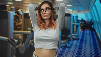 Fototapeta premium Woman with arms raised, hands behind head and exposed midriff in airport terminal, red hair and glasses wearing grey crop top and casual pose; playful ease.
