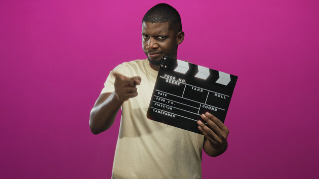 Young black man holding clapperboard and pointing finger at camera in studio; confidence assertiveness.