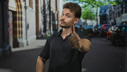 Naklejka premium Man pointing to chest on city street while giving thumbs up and looking at camera near parked cars and buildings, outdoors; confidence approval.