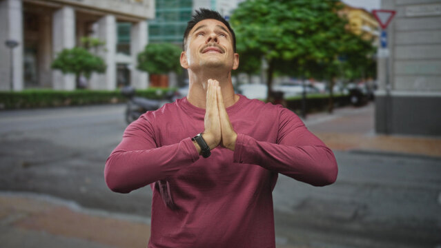 Man pressing hands in prayer gesture on a city street in front of a building, head tilted up showing moustache; gratitude reflection contemplation.