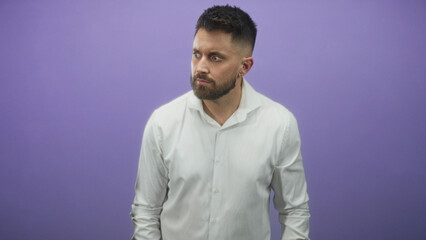 Obraz premium Man in white shirt pointing in studio against a purple backdrop, young and handsome with visible beard; doubt hesitation warning.