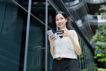 Fototapeta premium A young woman smiles while using her smartphone and holding a credit card outdoors near modern buildings.