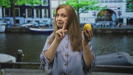 Fototapeta premium Woman holding lemon in left hand and points finger with right hand on a street by a canal; skepticism resilience.