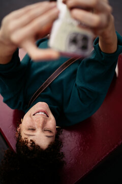 Smiling young woman with curly hair lies on red bench, holding up smartphone for a selfie, eyes closed in genuine joy. Captures a peaceful, personal digital moment in motion.