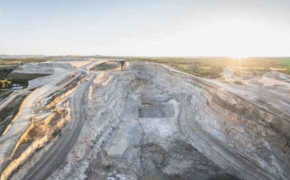Aerial view of a vast open-pit quarry with terraced excavation walls and winding dirt roads under a bright sun in Santa Lucia de Gordon, Castile and Leon, Spain.