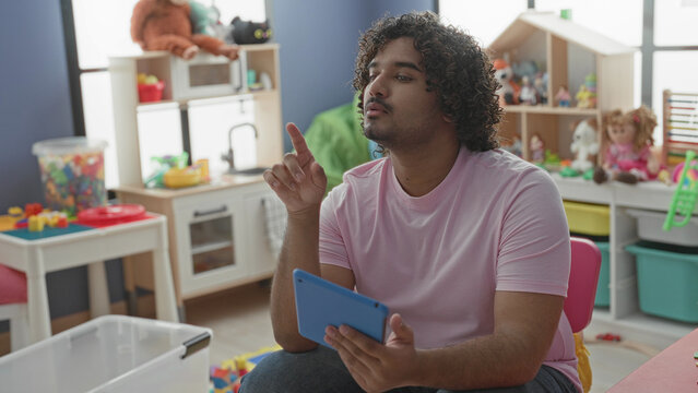 Man holding tablet and pointing finger while sitting on pink chair in school building playroom; quiet concentration teaching.