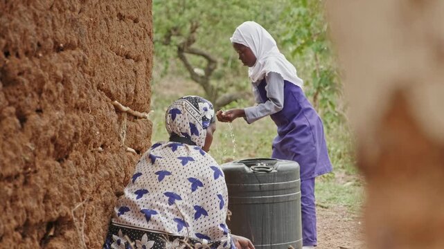 Back view of a woman washing dishes beside a mud house in rural Africa. The scene highlights water scarcity, limited resources, and the essential role of water in everyday survival and hygiene.