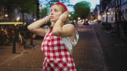 Fototapeta premium Woman chef in red check apron covering ears with hands on a dim cobblestone street lined with bicycles and lit shopfronts; distress noise.