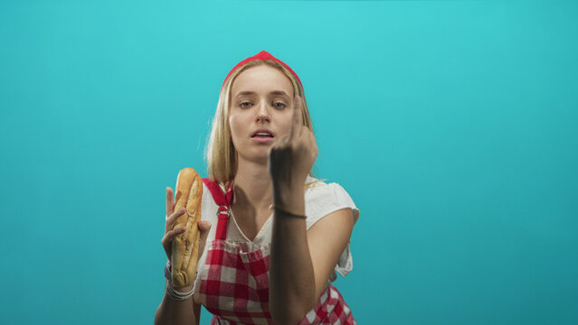 Woman chef in apron holds a baguette and flips her middle finger toward camera in studio; defiance rebellion.