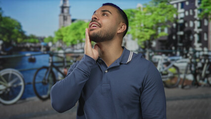 Fototapeta premium Young hispanic man places hand on chin next to canal railing with bicycles and urban buildings visible; contemplation.