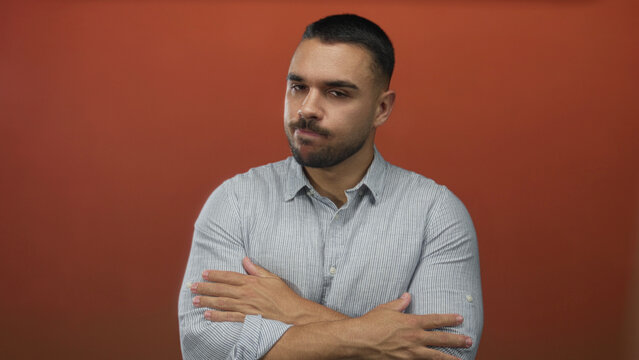 Hispanic man wearing striped shirt crosses arms in orange studio while closing eyes; rejection introspection.