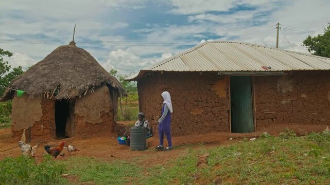 A young girl uses water near a mud house while a woman does chores in a rural African village. The scene highlights daily life, limited water resources, and the importance of water in basic routines.