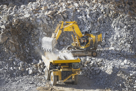 Aerial view of a large yellow excavator loading rocks into a massive dump truck in an open-pit mine surrounded by piles of stone debris in Santa Lucia de Gordon, Castile and Leon, Spain.