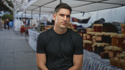 Obraz premium Man sitting at street market stall amid rows of preserve jars and striped tablecloth, facing camera; quiet contemplation.