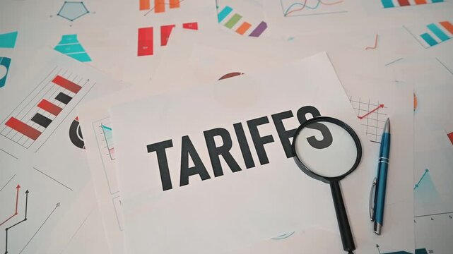 Documents related to tariffs are placed on a table filled with charts and graphs. A magnifying glass is shown focusing on the word tariffs indicating an analysis of economic factors