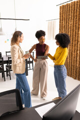 Fototapeta premium Diverse female colleagues standing at office holding tablet and travel mug, sparking discussion