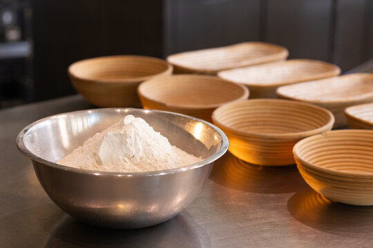 Stainless-steel mixing bowl holding mound of flour sitting left on countertop, bannetons visible